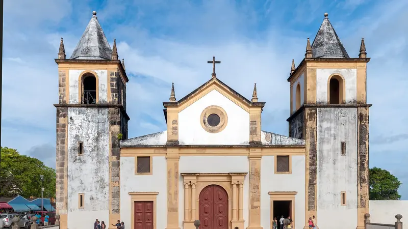 Fachada da Catedral da Sé de Olinda, com duas torres e paredes brancas e ocres sob céu azul, e pessoas na entrada